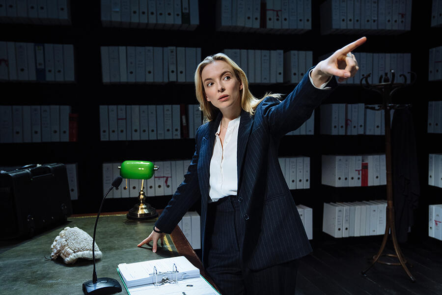 Jodie Comer behind a wooden desk, pointing at the audience.